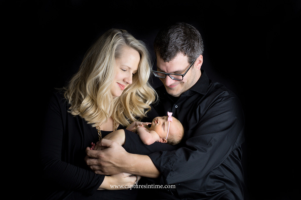 kansas city newborn baby photography mom and dad hold their newborn baby girl with a pink bow on her head on black backdrop