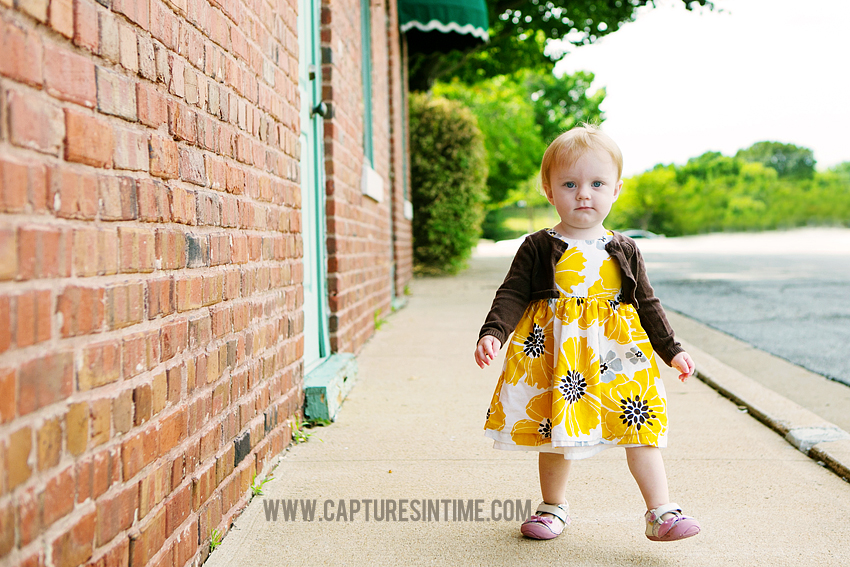 one year old girl floral dress walking next to brick building blue springs